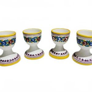 May include: Four white ceramic egg cups with floral designs. Each cup has a yellow rim and base, with a purple and white dotted pattern on the base. The floral design includes blue, red, and green accents.