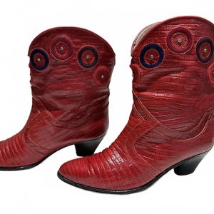 May include: A pair of red cowboy boots with a textured, reptile-like pattern. The boots have decorative circular accents in shades of red and blue on the upper shaft. The heels are black.