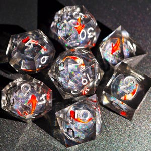 May include: Set of seven clear resin dice with a koi fish design. Each die features a red and white koi fish suspended within the transparent resin, with silver glitter and white numbering. The dice are designed for tabletop games.