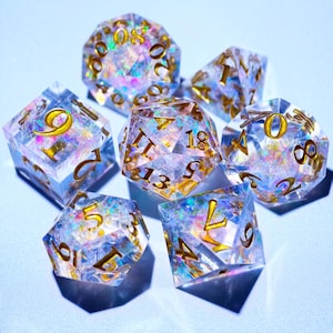 May include: Set of seven clear, multi-sided dice with gold-colored numbers. The dice have iridescent inclusions that reflect light. The dice are designed for tabletop games and role-playing games.