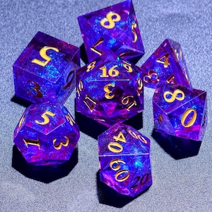 Puede incluir: Un conjunto de siete dados poliédricos en tonos morados y azules con números dorados. Los dados están hechos de un material translúcido, con un efecto arremolinado, similar a una galaxia. Para juegos de mesa.