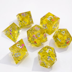 Può includere: Set di sette dadi in resina gialla con decorazioni floreali rosa e bianche e numeri argentati. I dadi hanno varie forme geometriche, tra cui d4, d6, d8, d10, d12 e d20, perfetti per i giochi da tavolo.