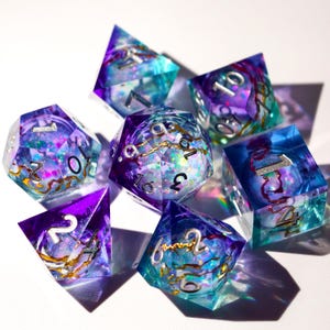 Può includere: Set di sette dadi poliedrici a tema fantasy. I dadi sono realizzati in resina trasparente con un mix di blu, viola e glitter iridescenti. I numeri sono argentati e all'interno ci sono vortici color oro.