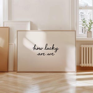 以下が含まれることがあります： 「how lucky are we」という言葉が黒い筆記体で書かれた額入りのプリント。プリントはクリーム色の背景と明るい木製のフレームです。他の額入りのアート作品が背景に見えます。