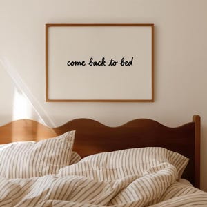 Puede incluir: Un cuadro enmarcado con las palabras "come back to bed" en escritura negra. El marco es marrón, y el cuadro está colgado sobre una cama con ropa de cama a rayas y un cabecero de madera.
