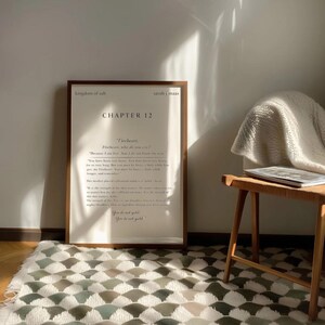 May include: A framed print with text from "Kingdom of Ash" by Sarah J. Maas, Chapter 12, in a wooden frame. The print is leaning against a white wall, with a wooden stool and patterned rug in the foreground. The text includes the words "Fireheart" and "You do not quit."