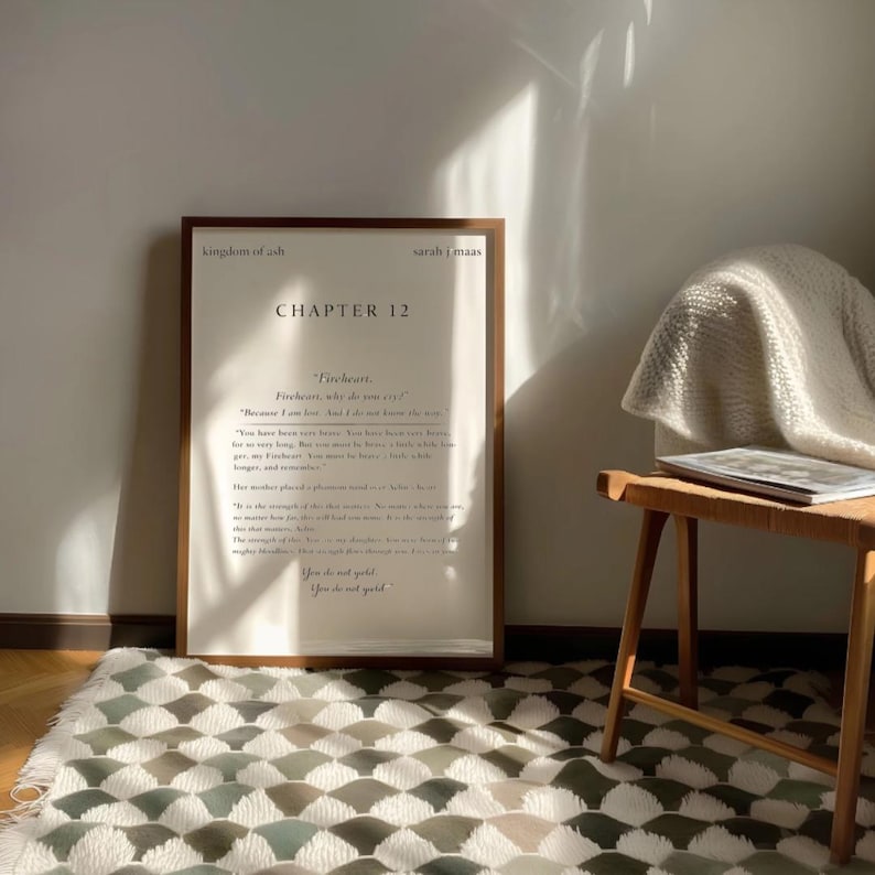 May include: A framed print with text from "Kingdom of Ash" by Sarah J. Maas, Chapter 12, in a wooden frame. The print is leaning against a white wall, with a wooden stool and patterned rug in the foreground. The text includes the words "Fireheart" and "You do not quit."