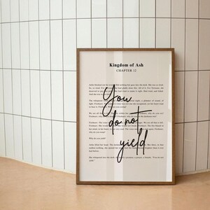 Può includere: Una stampa incorniciata con una cornice in legno. La stampa presenta il testo di "Kingdom of Ash, Capitolo 12". Le parole "You do not yield" sono scritte in corsivo sopra il testo stampato.