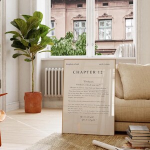 May include: A framed print with the text "kingdom of ash" and "CHAPTER 12" in black font. The print is leaning against a beige sofa. A potted plant with large green leaves is to the left of the print.
