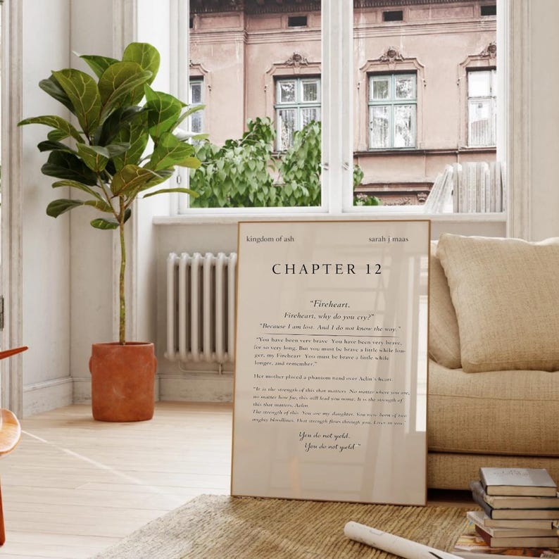 May include: A framed print with the text "kingdom of ash" and "CHAPTER 12" in black font. The print is leaning against a beige sofa. A potted plant with large green leaves is to the left of the print.