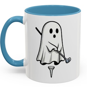 May include: White ceramic mug with a blue interior and handle. The mug features a cartoon ghost playing golf, with a golf club and tee. The ghost is white with black eyes and a black mouth. A fun gift for golfers.