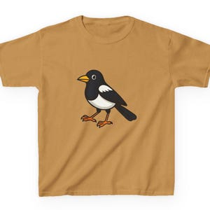 May include: Mustard-coloured t-shirt featuring a cartoon magpie. The bird is black and white with an orange beak and feet. The shirt is a short-sleeved crew neck, suitable for casual wear.