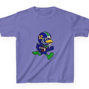 May include: A lilac-colored t-shirt featuring a cartoon bird wearing a football helmet with the number 12. The bird is running while holding a football. The helmet is blue with green and white stripes. The bird has a yellow beak and green feet.