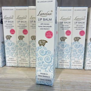 Puede incluir: Una caja rectangular blanca de bálsamo labial Lanolux con el texto NZ Lanolin Luxury, Lip Balm with manuka honey, bee propolis and vitamin E, y Made in New Zealand 15ml | 0.5oz. El envase muestra una ilustración de una oveja marrón.