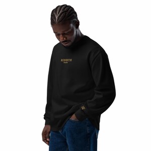 Peut inclure: Sweat-shirt noir à manches longues avec l'inscription dorée "REBIRTH Studio" sur la poitrine. Un logo doré est présent sur la manche. Le sweat-shirt est porté avec un jean bleu.