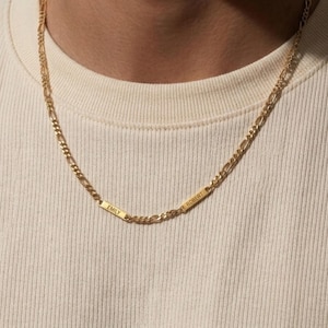 May include: A gold-colored chain necklace with two rectangular nameplates. One plate reads "EMILY" and the other reads "ROBERT". The necklace is worn around the neck and rests on a cream-colored shirt.