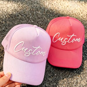 Puede incluir: Dos gorras de camionero, una rosa claro y otra rosa brillante, con la inscripción blanca "Custom". Las gorras tienen la parte trasera de malla y viseras curvas. Una mano sostiene la gorra rosa claro.