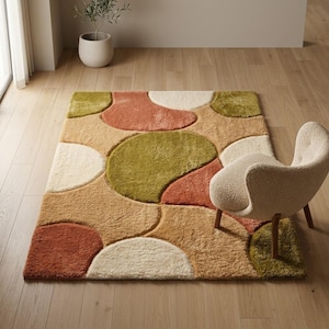 May include: A patterned rug featuring abstract shapes in beige, orange, green, and white. The rug is on a wooden floor, with a cream-colored chair and a potted plant visible in the background. The rug's dimensions are approximately 170 cm x 240 cm.