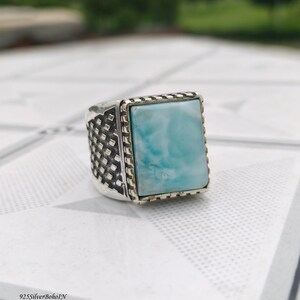 May include: A silver ring featuring a rectangular, light blue Larimar stone set in a bezel. The band has a dark, oxidized finish with a textured, grid-like pattern on the sides. The text 925SilverBohoIN is visible in the bottom left corner.