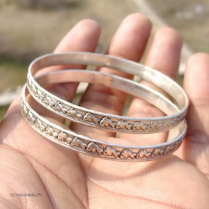 May include: Two silver bangles with an embossed elephant design. The bangles are round and have a slightly textured surface. The design is repeated around the entire circumference of the bangles. The bangles are held in a hand.