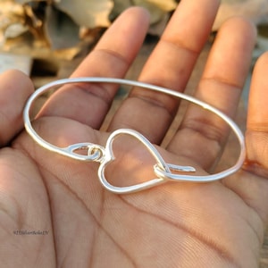 May include: A silver-colored bangle bracelet with a heart-shaped clasp. The bracelet is oval-shaped and appears to be made of a shiny metal. The heart clasp is a prominent design element. The text "925SilverBohoIN" is visible.