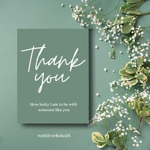 Op de afbeelding: Een saliegroene kaart met de woorden "Thank you" in witte letters. De kaart bevat ook de tekst "How lucky I am to be with someone like you". Witte bloemen en groen omringen de kaart.
