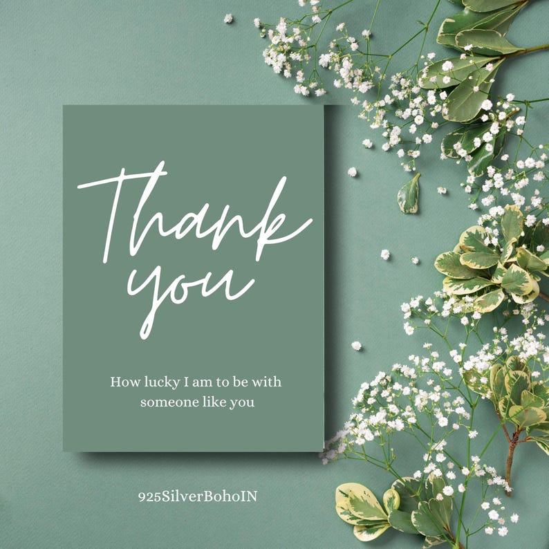 Op de afbeelding: Een saliegroene kaart met de woorden "Thank you" in witte letters. De kaart bevat ook de tekst "How lucky I am to be with someone like you". Witte bloemen en groen omringen de kaart.