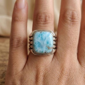 May include: A silver ring featuring a square, polished light blue Larimar gemstone with white marbling patterns. The metal band has a wide, tiered design with three horizontal ridges on each side of the central stone setting.