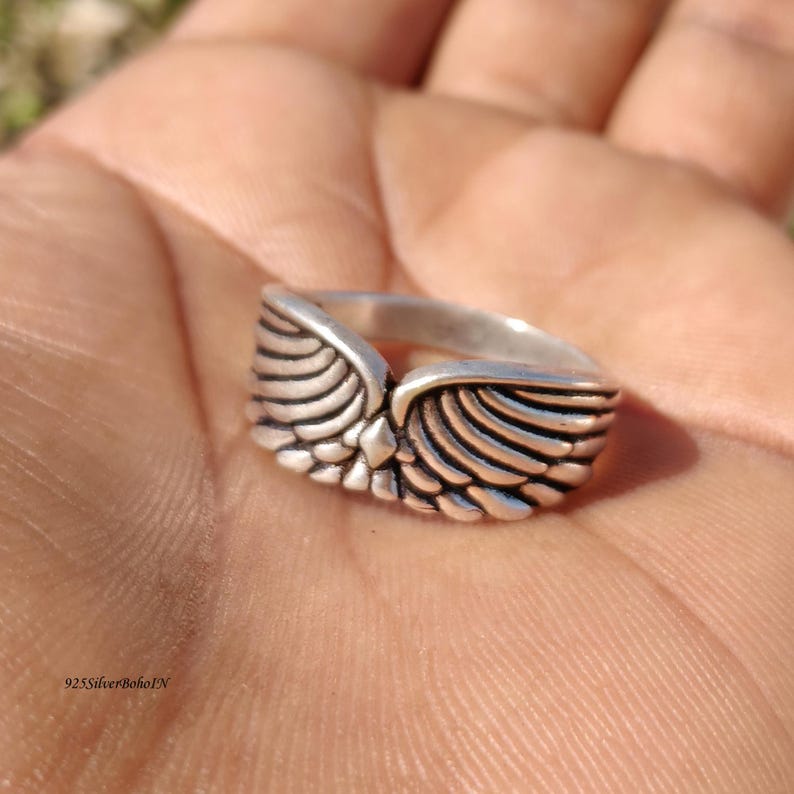 Pu&ograve; includere: Un anello d'argento con un design ad ali dettagliato. L'anello presenta un punto centrale dove le ali si incontrano, con intricati dettagli di piume. L'anello &egrave; mostrato su una mano, evidenziando il design e l'artigianato del gioiello.