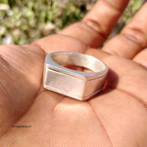 Può includere: Un anello d'argento con una faccia rettangolare e una fascia piatta. L'anello è tenuto in una mano, evidenziando la sua superficie lucida. Il design dell'anello è semplice e moderno. Il testo "925SilverBohoIN" è visibile.