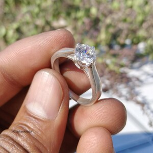 May include: A silver ring with a large, clear, round-cut gemstone. The ring is held in a hand, showcasing the bright, sparkling stone. The band is a simple, polished silver. The ring is a classic design, suitable for various occasions.
