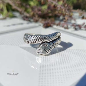Puede incluir: Un anillo plateado con forma de serpiente, con patrones de escamas detallados. La cabeza de la serpiente está en la parte delantera, y el cuerpo se enrolla alrededor del dedo. El anillo está sobre una superficie blanca con un fondo borroso.