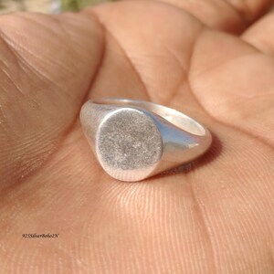 Puede incluir: Un anillo de sello plateado con una cara ovalada lisa. El anillo está hecho de un material metálico brillante y tiene un diseño simple y clásico. El anillo se muestra en un primer plano, destacando sus detalles.