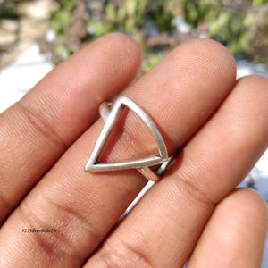 May include: A silver ring with a geometric triangle design. The ring features a hollow triangle shape, creating an open and modern look. The ring is shown on a finger, highlighting its size and style. The ring is a piece of jewelry.