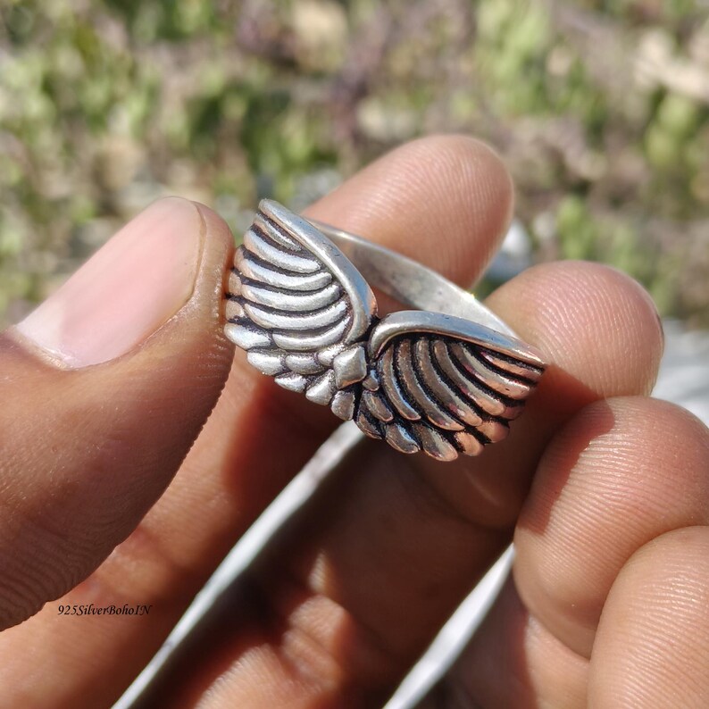 Pu&ograve; includere: Un anello color argento con un design ad ali dettagliato. L'anello presenta due ali che si incontrano al centro, ogni ala ha pi&ugrave; linee a forma di piuma. L'anello &egrave; tenuto in una mano.