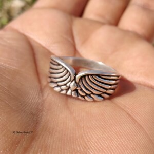 Pu&ograve; includere: Un anello d'argento con un design ad ali dettagliato. L'anello presenta un punto centrale dove le ali si incontrano, con intricati dettagli di piume. L'anello &egrave; mostrato su una mano, evidenziando il design e l'artigianato del gioiello.
