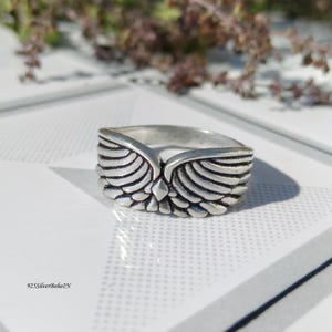 Pu&ograve; includere: Un anello d'argento con un design ad ali d'angelo. L'anello presenta dettagliate trame di piume e un accento centrale a forma di diamante. L'anello &egrave; di forma quadrata e presenta il testo "925SilverBohoIN" in basso a sinistra.