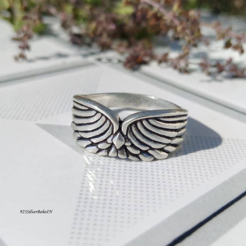 Pu&ograve; includere: Un anello d'argento con un design ad ali d'angelo. L'anello presenta dettagliate trame di piume e un accento centrale a forma di diamante. L'anello &egrave; di forma quadrata e presenta il testo "925SilverBohoIN" in basso a sinistra.