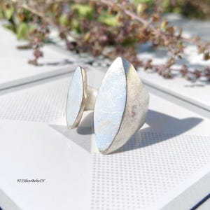 May include: A silver ring with a geometric design. The ring features two elongated, oval-shaped silver elements. The ring is set against a white surface with a subtle pattern, with a blurred background of foliage. The text "925SilverBohoIN" is visible.
