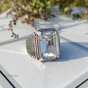Puede incluir: Un anillo de plata con una gema clara rectangular grande. El anillo tiene una banda texturizada y un engaste detallado alrededor de la piedra. El anillo se muestra sobre una superficie blanca.