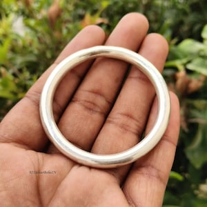 Puede incluir: Una pulsera de plata lisa y pulida con un perfil circular redondeado. La superficie metálica es reflectante y de grosor uniforme, formando un bucle continuo y sin costuras. El texto 925SilverBohoIN aparece impreso en la esquina inferior izquierda.