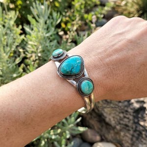 May include: A silver cuff bracelet featuring three turquoise stones. The central stone is large and pear-shaped, flanked by two smaller, round turquoise stones. Each stone is set in a silver bezel with a twisted rope border detail.