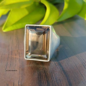 May include: A rectangular, smoky quartz gemstone ring with a silver band. The gemstone is a light brown colour and is set in a simple, modern design. The ring is displayed on a dark wooden surface with green leaves in the background.
