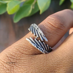 Op de afbeelding: Een zilverkleurige ring met een vleugelontwerp. De ring is open en wikkelt zich om de vinger, met gedetailleerde, veerachtige patronen. De ring wordt getoond op de vinger van een persoon, met een vage achtergrond van groene bladeren.
