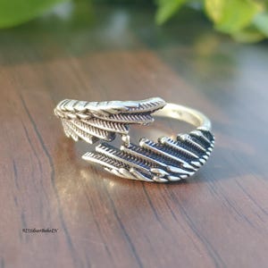 Op de afbeelding: Een zilveren ring met een veermotief. De ring heeft een gespleten schacht en gedetailleerde veersculpturen. Gepresenteerd op een houten oppervlak. De tekst "925SilverBohoIN" is zichtbaar.