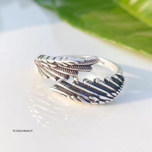 Op de afbeelding: Een zilverkleurige ring met een veermotief. De ring heeft twee gedetailleerde veerelementen die zich om de vinger wikkelen. De ring staat op een wit oppervlak met een groen blad op de achtergrond. De tekst "925SilverBohoIN" is zichtbaar.