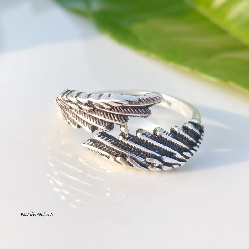 Op de afbeelding: Een zilverkleurige ring met een veermotief. De ring heeft twee gedetailleerde veerelementen die zich om de vinger wikkelen. De ring staat op een wit oppervlak met een groen blad op de achtergrond. De tekst "925SilverBohoIN" is zichtbaar.