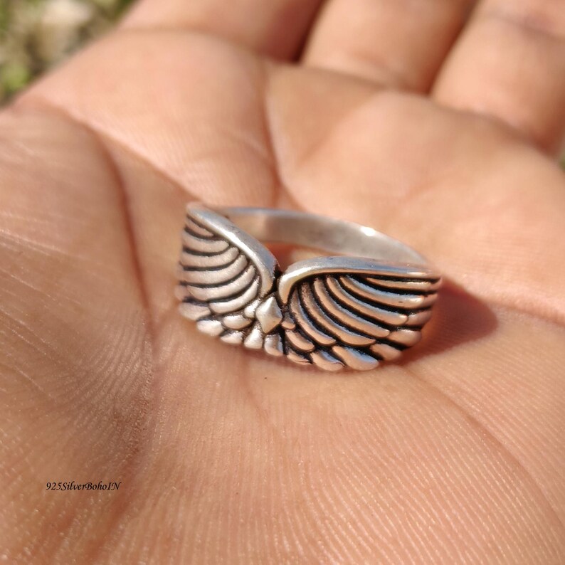 Pu&ograve; includere: Un anello d'argento con un design ad ali dettagliato. L'anello presenta un punto centrale dove le ali si incontrano, con intricati dettagli di piume. L'anello &egrave; mostrato in un primo piano, evidenziando la consistenza e il design del gioiello.