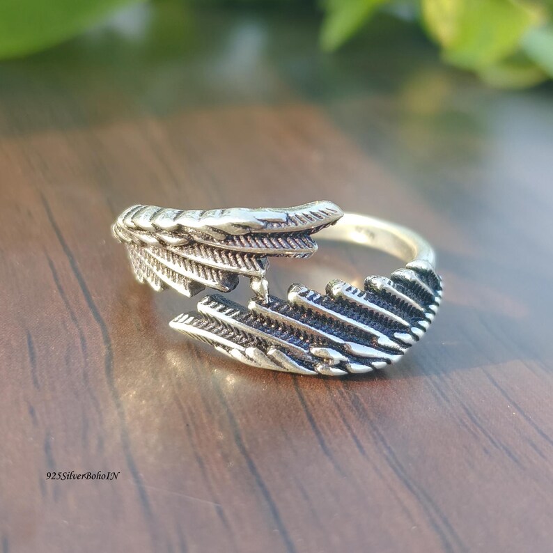 Op de afbeelding: Een zilverkleurige ring met een veermotief. De ring heeft een gespleten schacht en wikkelt zich om de vinger. De veerd details zijn ingewikkeld gesneden. De ring staat op een houten oppervlak. De tekst "925SilverBohoIN" is zichtbaar.