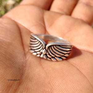 Pu&ograve; includere: Un anello d'argento con un design ad ali dettagliato. L'anello presenta un punto centrale dove le ali si incontrano, con intricati dettagli di piume. L'anello &egrave; mostrato in un primo piano, evidenziando l'artigianato e il design.